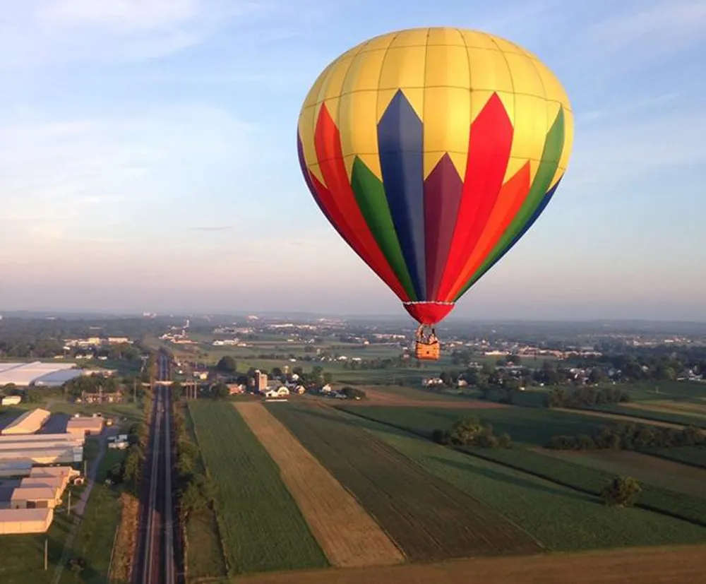 A colorful hot air balloon floats above a rural landscape with farmlands and a straight road in the early morning light