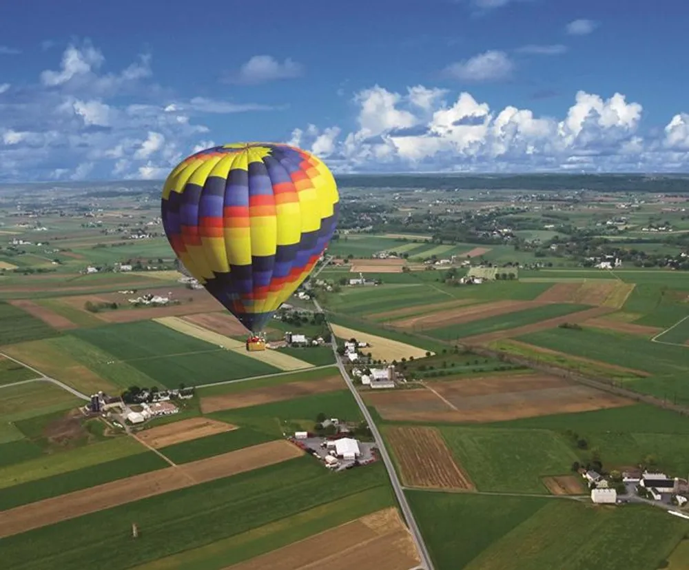 A colorful hot air balloon floats above a patchwork of farmland under a bright blue sky with scattered clouds