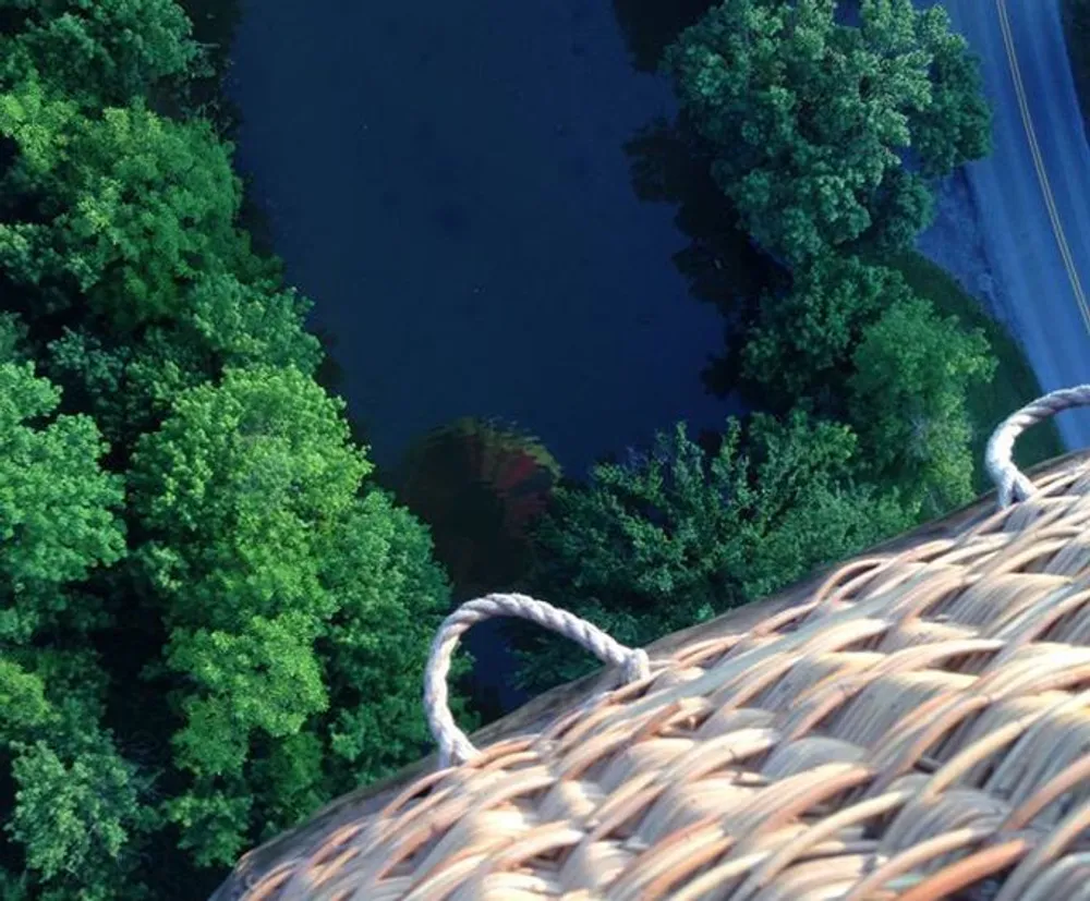 The image shows an aerial view from the edge of a hot air balloon basket looking down at the tops of trees and a reflective body of water