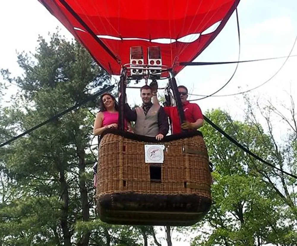Three people are standing in the basket of a red hot air balloon which appears to be on the ground or at low altitude amidst a green treed area