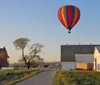 A colorful hot air balloon floats above a rural landscape with farmlands and a straight road in the early morning light