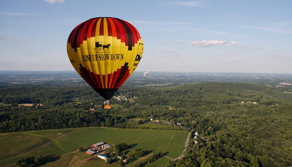 A hot air balloon adorned with the message COME UP SLOW DOWN is floating above a scenic landscape with green fields and a forest