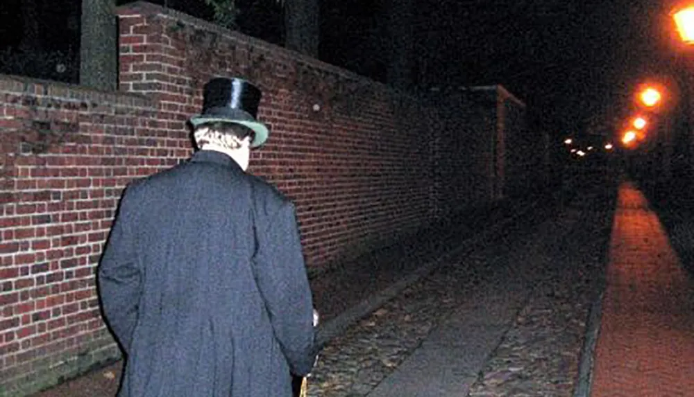 A person wearing a top hat and coat is walking along a brick-lined alleyway at night