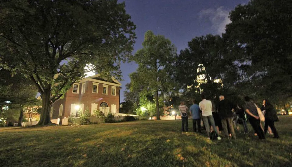 A group of people is gathering at night on a grassy area near an illuminated historical building with tall trees and a city building in the background