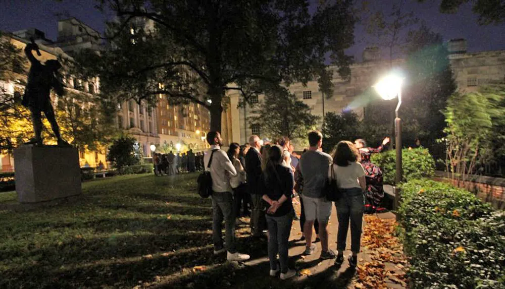 A group of people is gathered at night in an outdoor setting possibly listening to a speaker or guide near a statue and street light