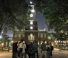 A group of people is standing at night in front of a historic building with a clock tower illuminated under the evening sky