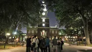 A group of people is standing at night in front of a historic building with a clock tower, illuminated under the evening sky.