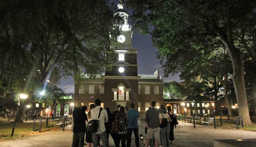 A group of people is standing at night in front of a historic building with a clock tower illuminated under the evening sky