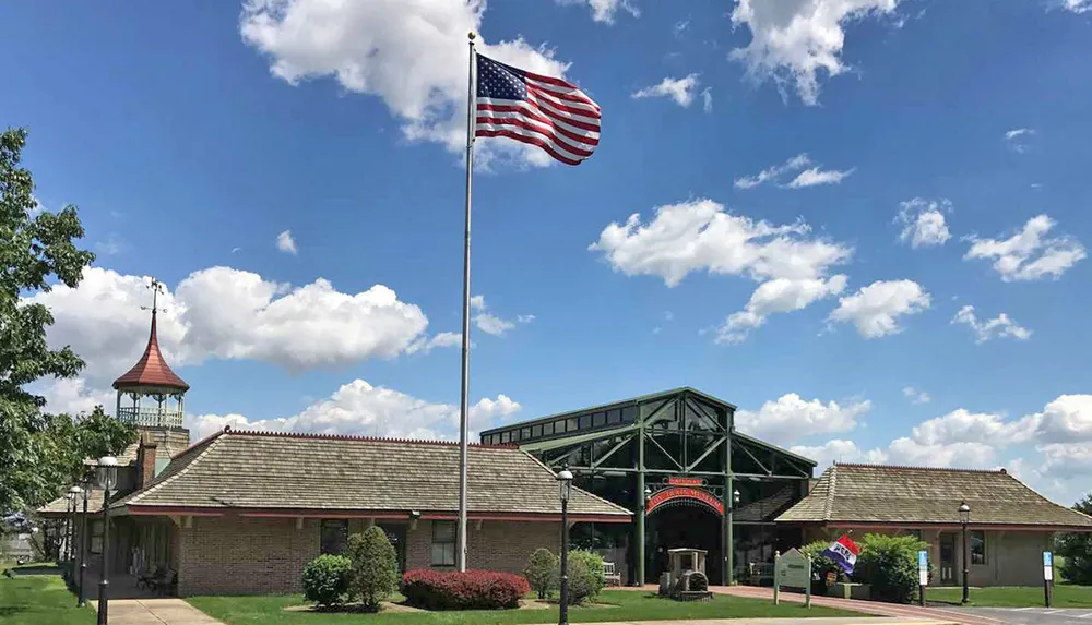 The photograph shows an American flag on a tall pole waving in front of a building with a distinctive clock tower and covered entrance set against a blue sky with scattered clouds