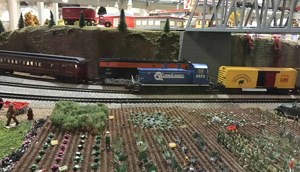 The image shows a detailed miniature model train setup with a blue and orange locomotive rolling stock a carefully crafted small-scale landscape with fields