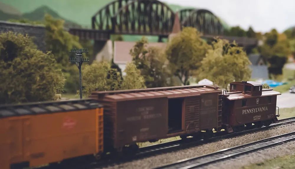 This image displays a detailed model train set featuring a Pennsylvania railroad caboose and freight cars set in a scenic miniature landscape with trees and a bridge in the background