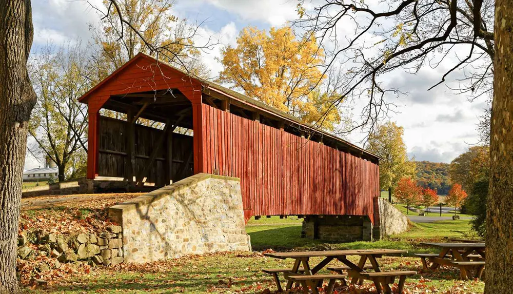 A red covered bridge spans across a stone base amidst a scenic autumn landscape with picnic tables in the foreground