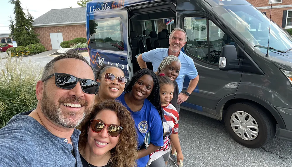 A diverse group of seven people are smiling for a selfie in front of a van with the text Live Life Fully on it