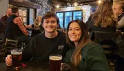 A smiling man and woman are sitting at a table with pints of beer in a cozy pub setting.