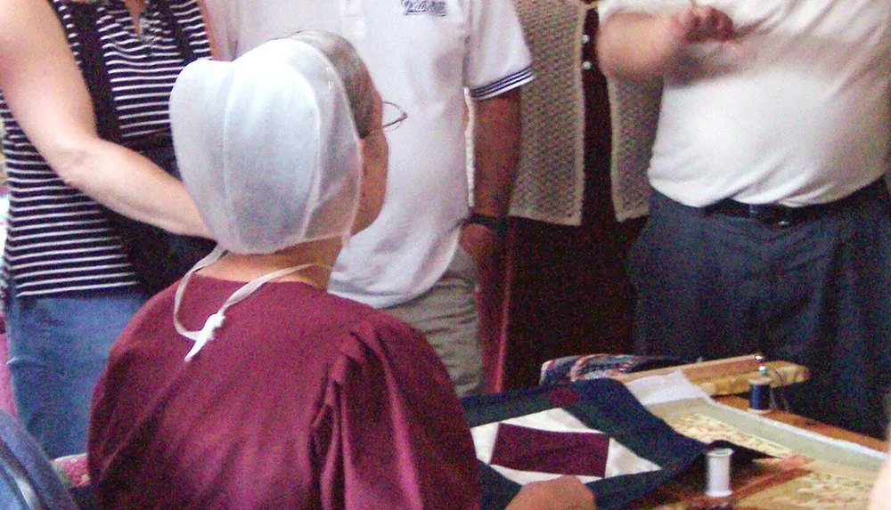 A person wearing a headscarf sits in front of a textile craft setup amidst a group of people in a room