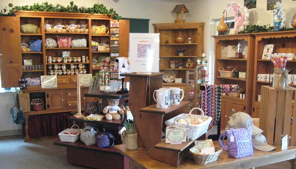The image shows a quaint and cozy gift shop interior filled with an assortment of colorful items ranging from quilts and pillows to ceramics and decorative objects displayed on wooden shelving and tables