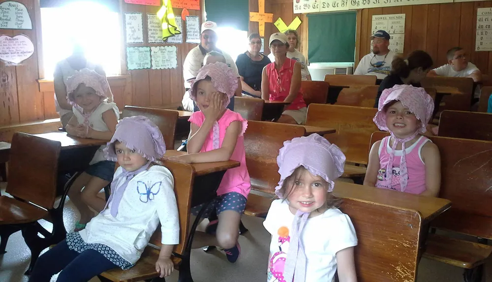Children and adults are sitting in an old-fashioned classroom with some of the youngsters wearing matching bonnets and smiling at the camera