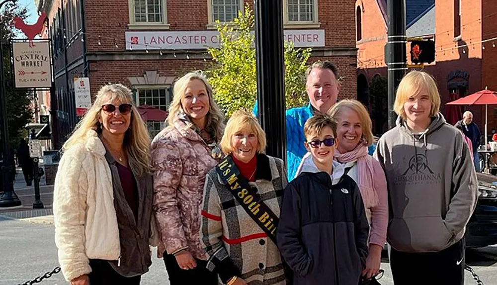 A group of seven people including a woman wearing a sash that says Its My 80th Birthday are smiling for a photo outdoors with a building labeled Lancaster Central Market in the background
