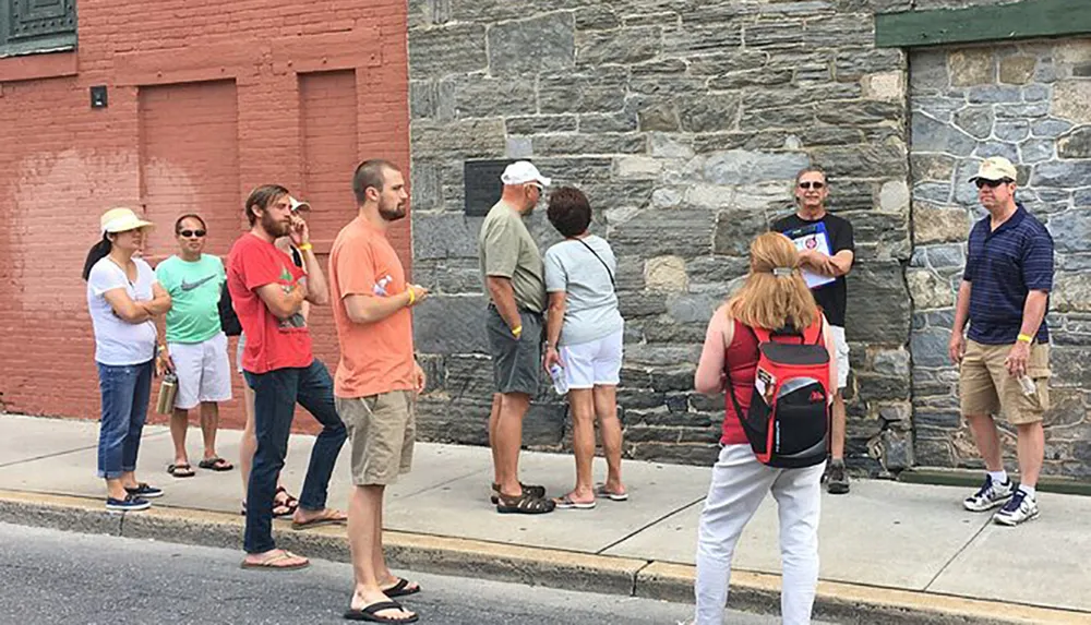A group of people are standing outdoors on a sidewalk seemingly listening or waiting with some engaged in conversation and others looking in various directions
