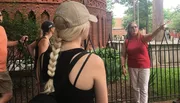 A group of people is listening to a woman who appears to be giving a tour or presentation outdoors.