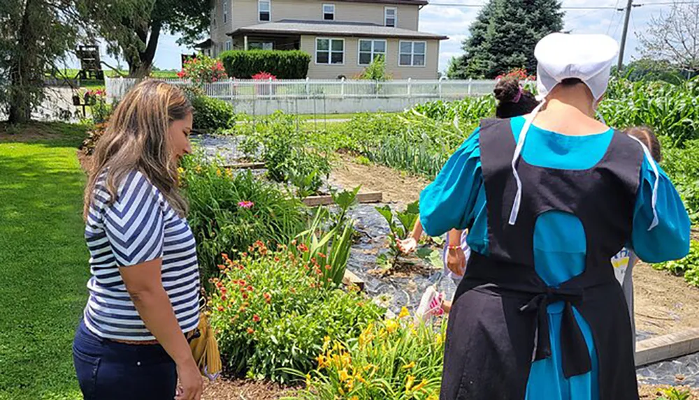 Two individuals one in traditional Amish clothing are standing near a garden and conversing in a sunny rural setting