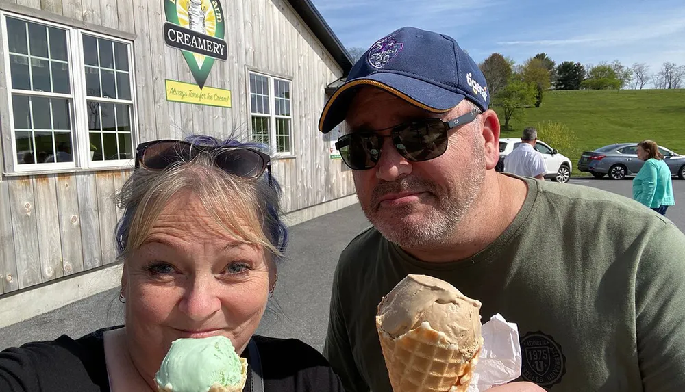 Two people are taking a selfie outside a creamery each holding an ice cream cone and smiling on a sunny day