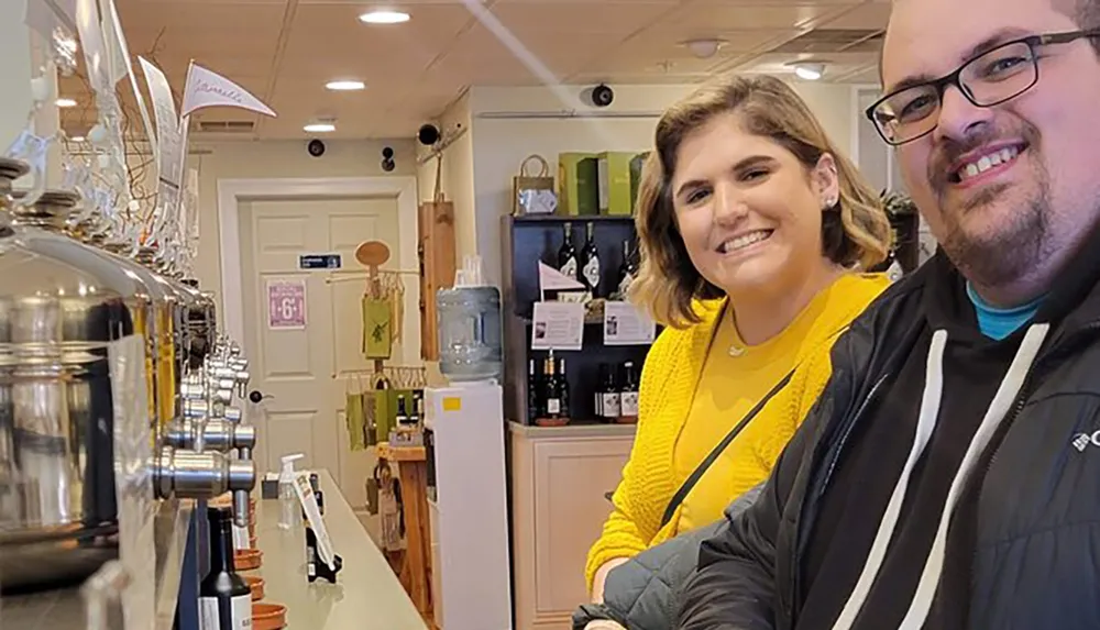 Two smiling people pose for a selfie inside a cozy shop with dispensers and various items displayed on the shelves in the background