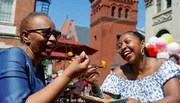 Two women are laughing joyfully while sharing a meal outdoors on a sunny day.