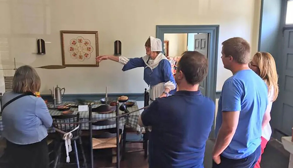A woman in historical clothing is explaining something to a group of attentive visitors in a room with old-fashioned cookware