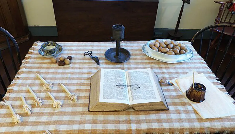 The image depicts a rustic historical setting with a large open book and spectacles in the center of a checkered tablecloth flanked by items like candles nuts and a metal candle holder suggesting an old-fashioned domestic scene or a historical reenactment display
