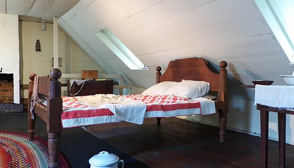 The image shows a rustic attic bedroom with a traditional wooden bed quilted cover and simple furnishings including a bedside table with a chamber pot underneath conveying a sense of historical domestic life