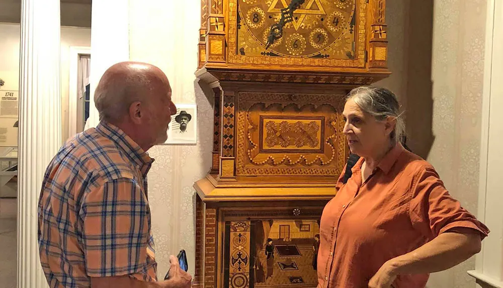 A man and a woman are looking at an ornately decorated antique clock in a room with vintage wallpaper