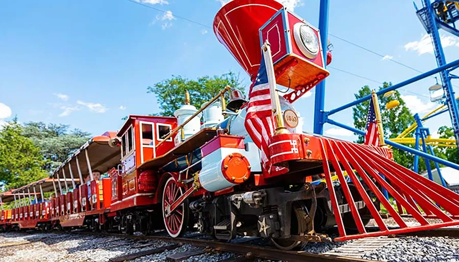 The image shows a brightly colored red train with flags, set against a backdrop of blue skies and a partially visible amusement park.