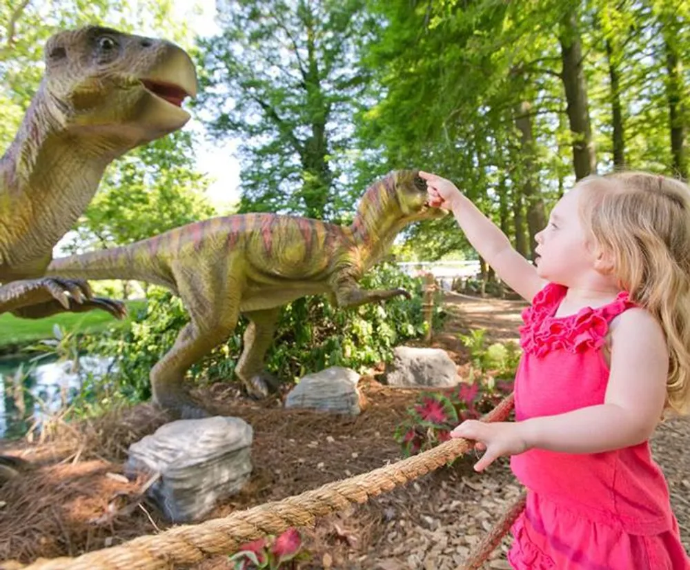 A young girl in a pink dress appears enchanted as she reaches out towards a lifelike model of a dinosaur in a lush garden setting