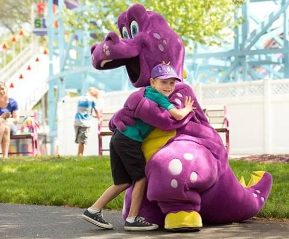A child is joyfully hugging a person in a purple dinosaur costume at what appears to be an outdoor event or amusement park