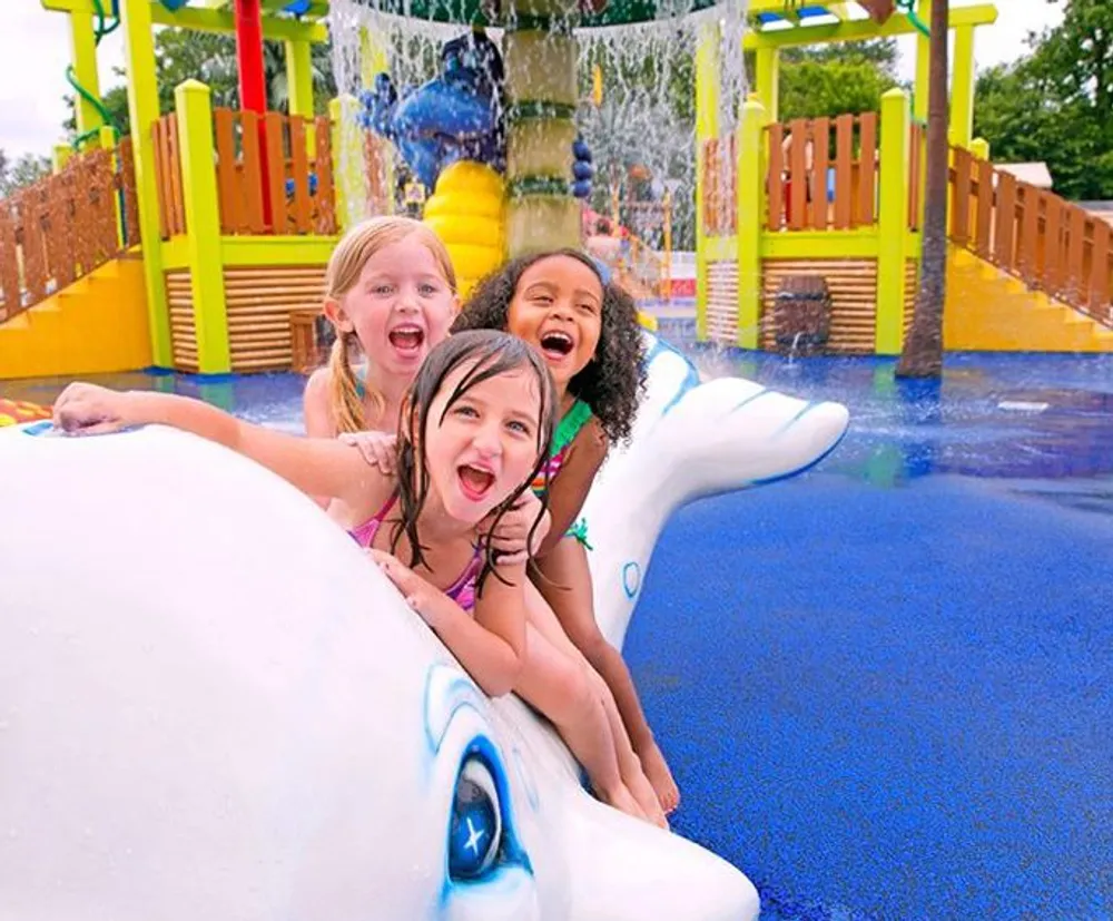 Three children are gleefully playing on a water park slide that resembles a white dolphin with a colorful water play structure in the background