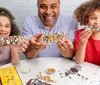 A man and two children are smiling while showing off their personally decorated chocolate bars at a table scattered with various candy toppings