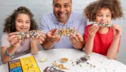 A man and two children are smiling while showing off their personally decorated chocolate bars at a table scattered with various candy toppings.
