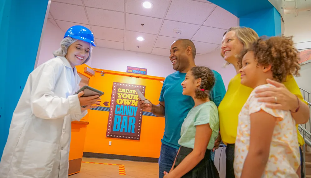 A smiling family is interacting with a worker in a protective suit and hard hat near a Create Your Own Candy Bar station
