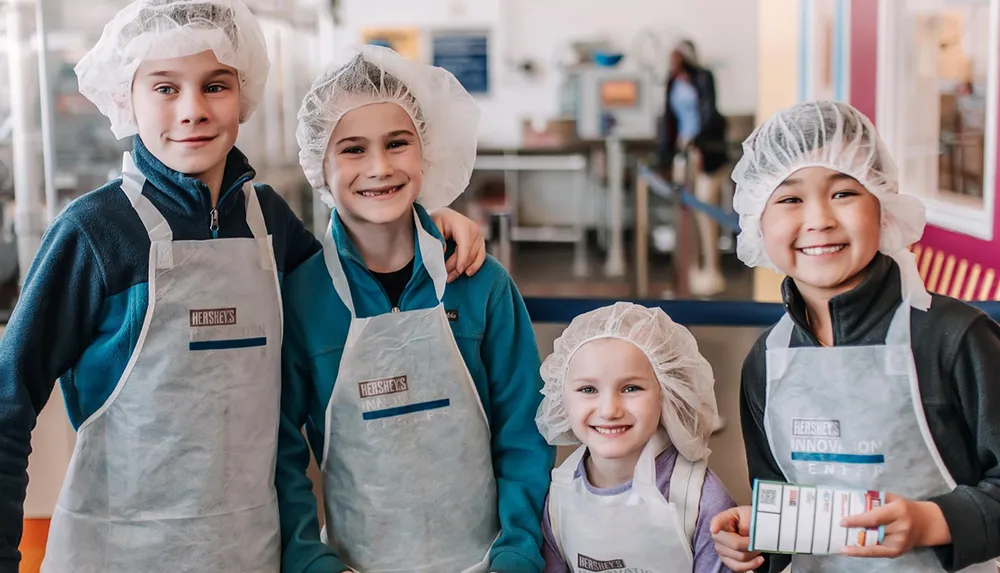 A group of smiling children are wearing hairnets and aprons branded with Hersheys Chocolate World suggesting they are participating in a chocolate-making activity or tour