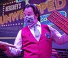 An individual in a steampunk-inspired costume holds up a large whimsical cheese wedge prop on stage entertaining an audience that includes both focused and distracted members