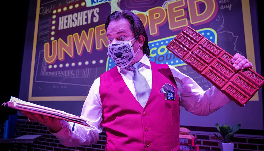 A person wearing a vest and a face mask with Hershey's branding holds an oversized Hershey's chocolate bar prop in a colorful setting featuring Hershey's Unwrapped in the background.