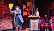 An individual in a steampunk-inspired costume holds up a large, whimsical cheese wedge prop on stage, entertaining an audience that includes both focused and distracted members.