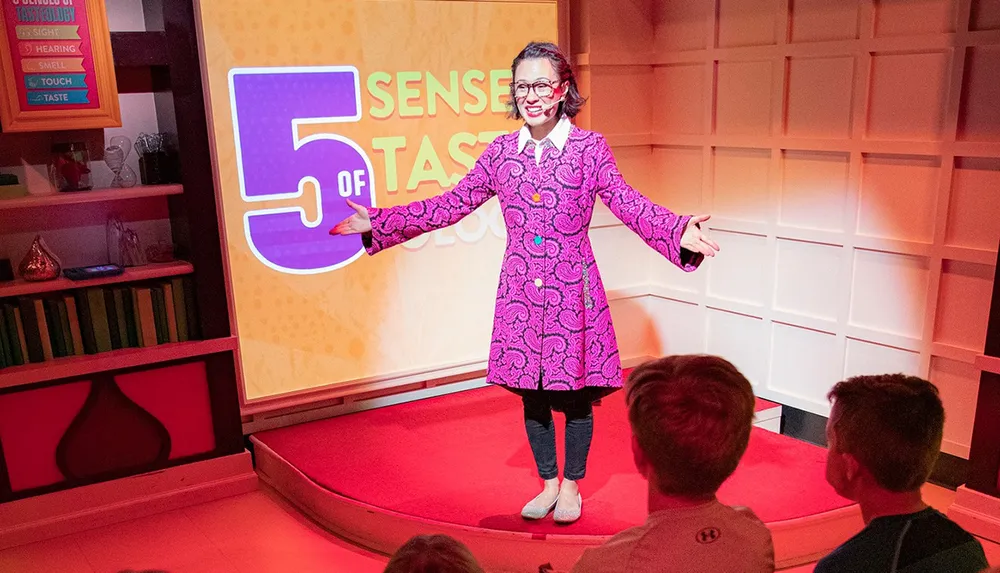 A woman is presenting on a stage with the text 5 SENSES OF TASTE behind her while children in the audience watch attentively