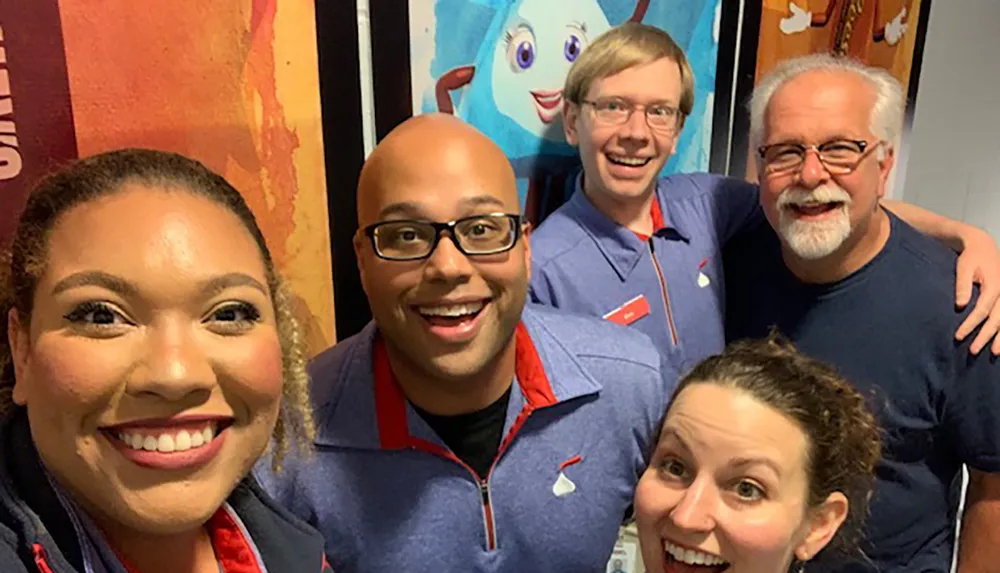 A group of five people are smiling for a selfie with colorful animated characters depicted on a poster in the background