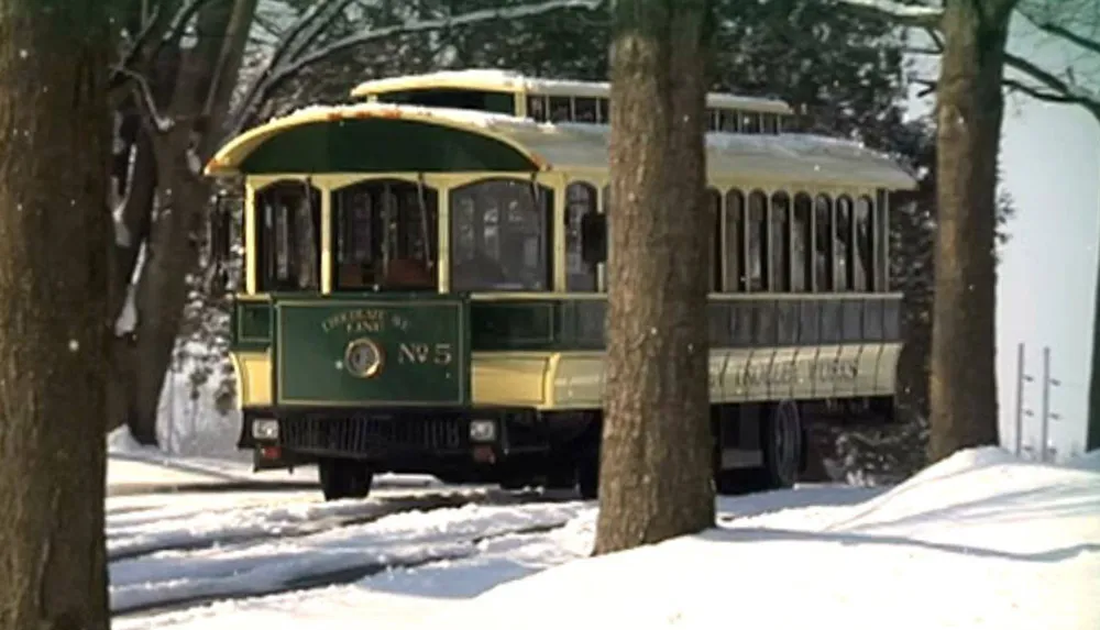 A vintage green and yellow tram is traveling through a snow-covered landscape flanked by trees