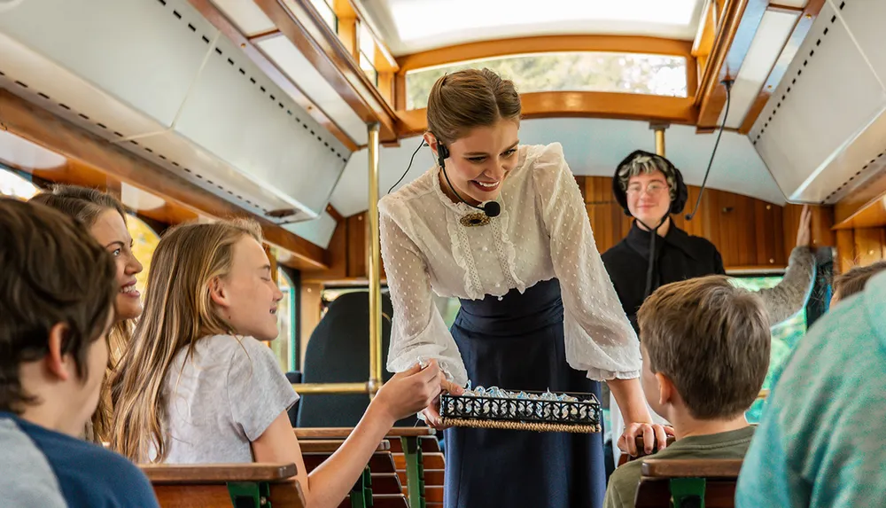 A cheerful woman in historical attire is engaging with passengers likely performing or narrating in a vintage train carriage