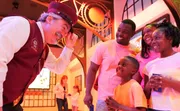 A performer in a conductor's outfit is entertaining a smiling family at what appears to be a themed entertainment venue or event.
