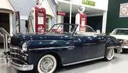 A classic convertible car is on display in a retro-themed exhibit with vintage gas pumps and signage in the background.