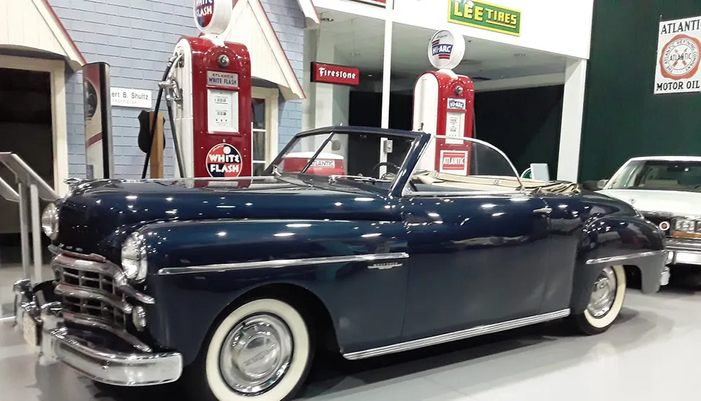A classic convertible car is on display in a retro-themed exhibit with vintage gas pumps and signage in the background